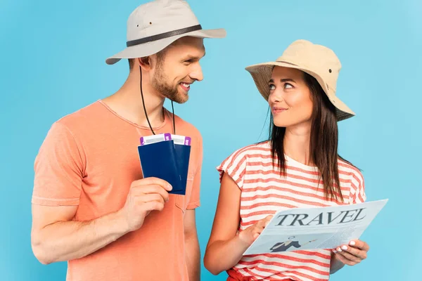 Glückliches Mädchen mit Hut hält Reisezeitung in der Hand und schaut Mann mit Pässen isoliert auf blauem Grund an — Stockfoto