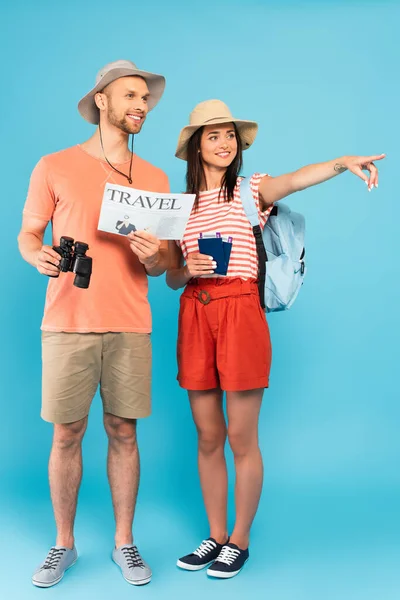 Happy girl in hat pointing with finger and holding passports near man with travel newspaper and binoculars on blue — Stock Photo