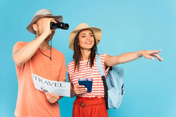 Ragazza felice indicando con il dito e tenendo i passaporti vicino all'uomo con giornale di viaggio guardando attraverso binocoli isolati su blu — Foto stock