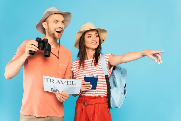Chica feliz señalando con el dedo y sosteniendo pasaportes cerca hombre emocionado con periódico de viaje y prismáticos aislados en azul - foto de stock