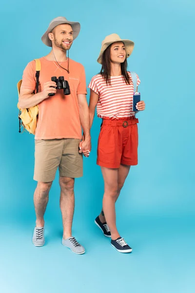 Chica feliz con pasaportes tomados de la mano con novio alegre caminando sobre azul - foto de stock