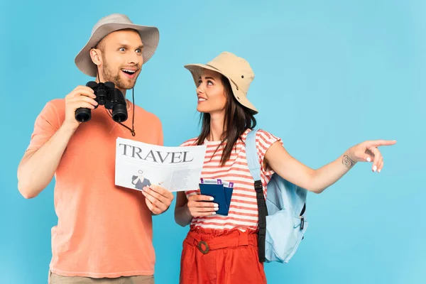 Щаслива жінка вказує пальцем і тримає паспорти біля збудженого чоловіка з туристичною газетою і біноклями ізольовані на синьому — стокове фото