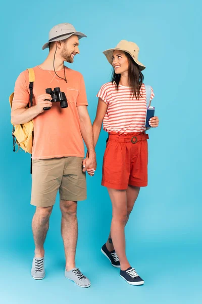 Viajeros felices en sombreros mirándose unos a otros y tomándose de la mano mientras caminan sobre azul - foto de stock