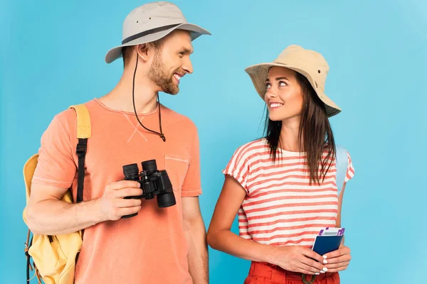 Happy travelers in hats looking at each other while holding passports and binoculars isolated on blue — Stock Photo