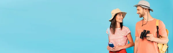 Imagem horizontal de viajantes felizes em chapéus olhando uns para os outros enquanto segurando passaportes e binóculos isolados em azul — Fotografia de Stock