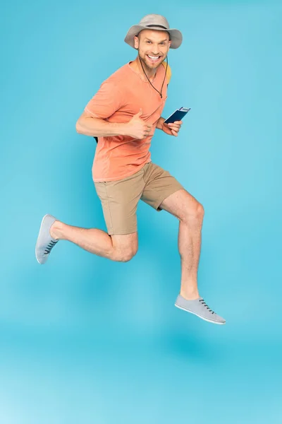 Cheerful man in hat holding passport, jumping and showing thumb up on blue — Stock Photo