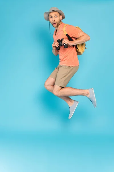Schockierter Mann mit Hut hält Fernglas in der Hand und springt auf blau — Stockfoto