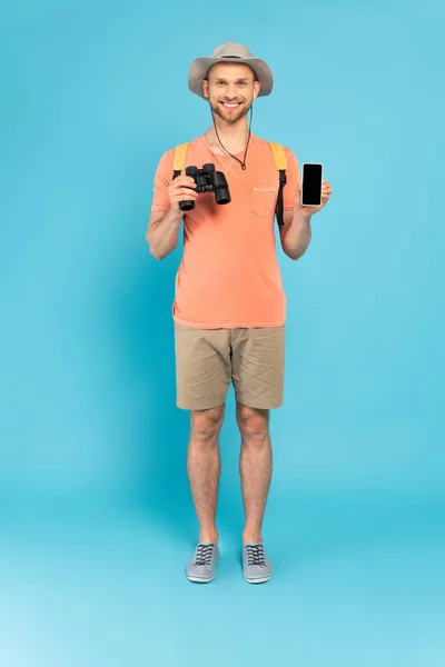 Hombre feliz sosteniendo binoculares y teléfono inteligente con pantalla en blanco en azul - foto de stock