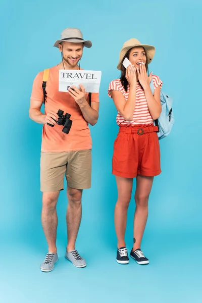 Felice uomo leggendo giornale di viaggio vicino ragazza in cappello parlando su smartphone su blu — Foto stock