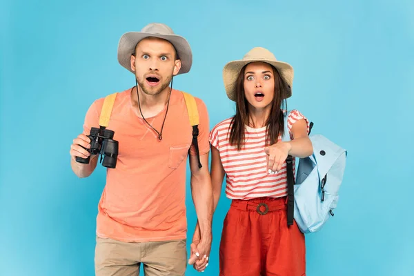 Shocked woman in hat pointing with finger near boyfriend with binoculars isolated on blue — Stock Photo