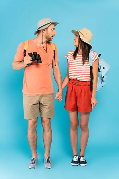 Потрясенная пара в шляпах, держащихся за руки и смотрящих друг на друга в голубом — стоковое фото