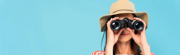 Colpo panoramico di giovane donna in cappello guardando attraverso binocoli isolati su blu — Foto stock
