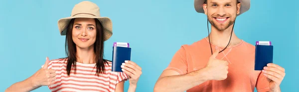 Panoramic shot of happy man and woman in hats holding passports and showing thumbs up isolated on blue — Stock Photo