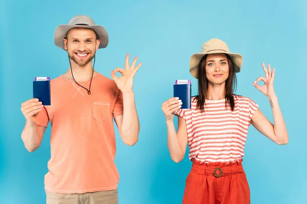 Happy man and woman in hats holding passports and showing ok sign on blue — Stock Photo