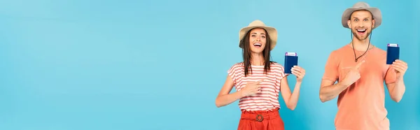 Panoramic crop of happy man and woman in hats pointing with fingers at passports isolated on blue — Stock Photo