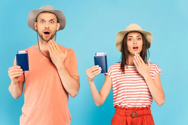 Sorprendido hombre y mujer en sombreros mirando a la cámara y sosteniendo pasaportes en azul - foto de stock