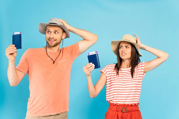 Confused man and woman touching hats and holding passports on blue — Stock Photo