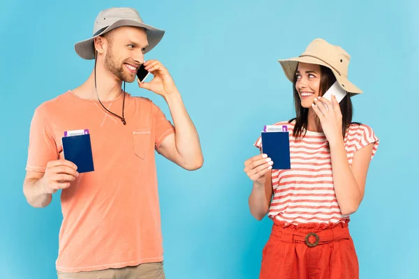 Happy couple in hats talking on smartphones, holding passports and looking at each other isolated on blue — Stock Photo