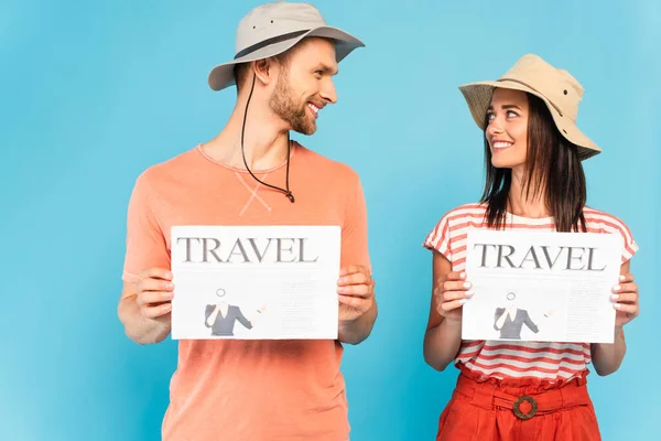 Happy couple in hats holding travel newspapers and looking at each other on blue — Stock Photo