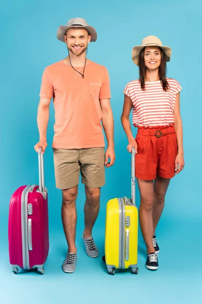 Glückliches Paar in Hüten, die neben Reisetaschen auf blau stehen — Stockfoto