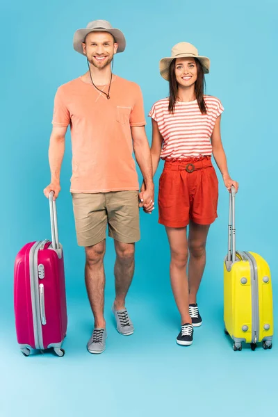 Feliz pareja en sombreros de pie cerca de bolsas de viaje y tomados de la mano en azul - foto de stock