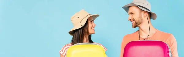 Plano panorámico de feliz pareja mirándose unos a otros y sosteniendo el equipaje aislado en azul - foto de stock