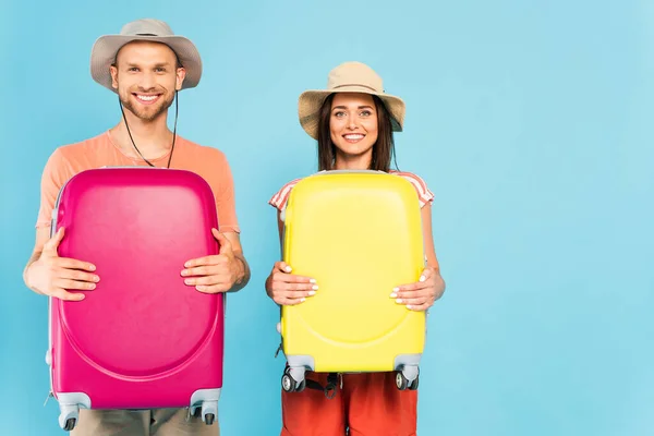 Glückliches Paar blickt in die Kamera und hält Gepäck isoliert auf blauem Grund — Stockfoto