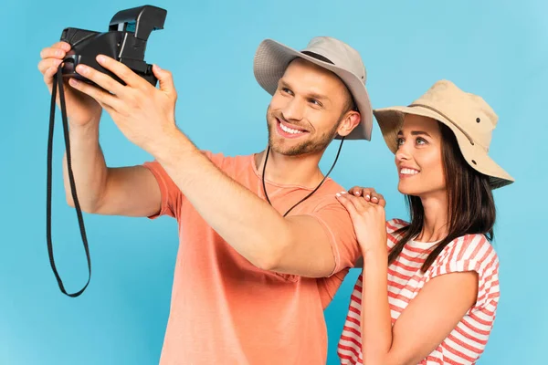 Felice uomo in cappello tenendo fotocamera vintage e prendendo selfie con allegra ragazza isolata su blu — Foto stock