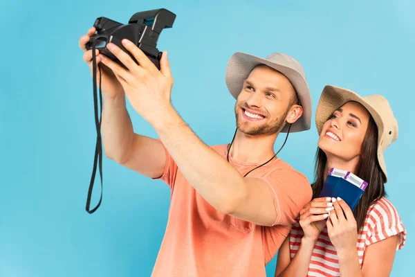 Felice uomo in cappello tenendo fotocamera vintage e prendendo selfie con allegra ragazza isolata su blu — Foto stock