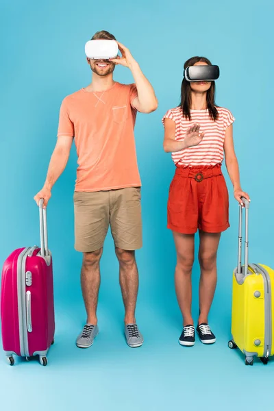 Счастливая пара в наушниках виртуальной реальности, стоящая с багажом на синем — стоковое фото