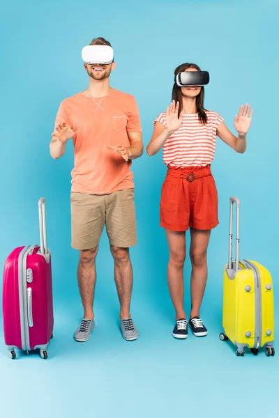 Cheerful couple in virtual reality headsets standing with luggage on blue — Stock Photo