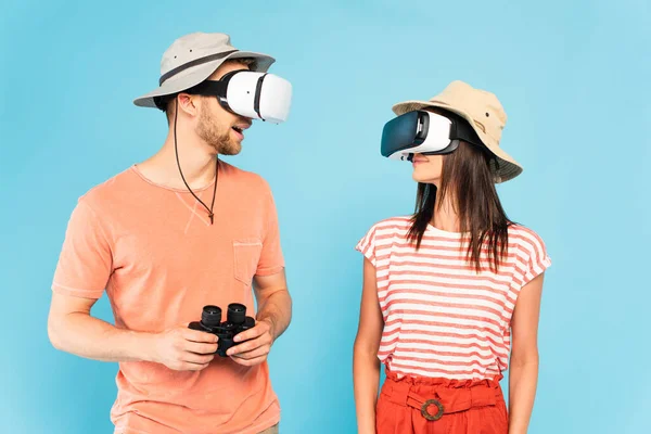 Sorprendido hombre en realidad virtual auricular celebración de prismáticos cerca de la mujer aislada en azul - foto de stock