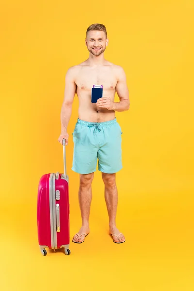 Щасливий і м'язистий чоловік тримає паспорт і стоїть з багажем на жовтому — стокове фото