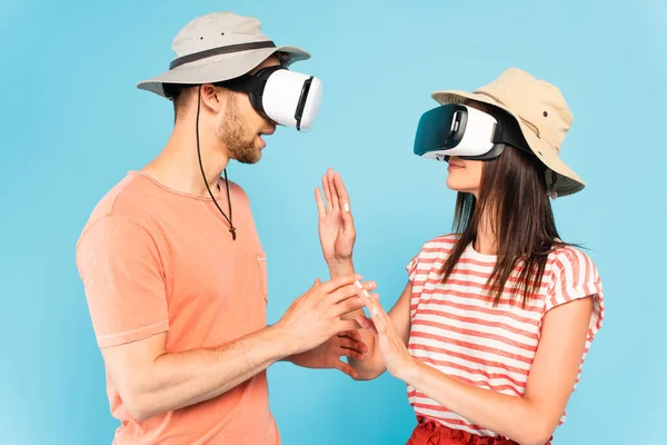 Couple in hats and virtual reality headsets gesturing isolated on blue — Stock Photo