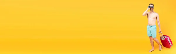 Raccolto panoramico di uomo felice guardando attraverso binocoli e camminando con bagaglio su giallo — Foto stock