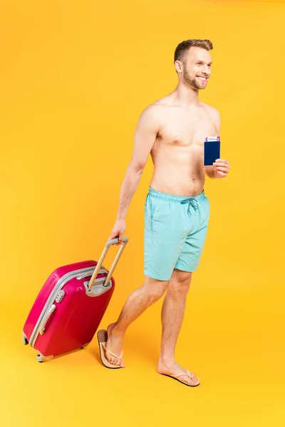 Щасливий і мускулистий чоловік тримає паспорт і ходить з багажем на жовтому — стокове фото