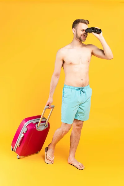 Happy man looking through binoculars and standing with baggage on yellow — Stock Photo