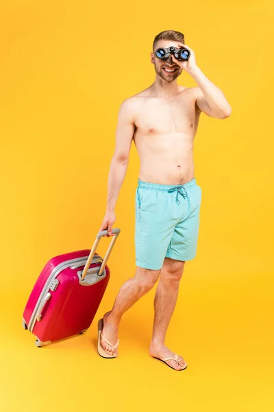Щасливий м'язистий чоловік дивиться через бінокль і стоїть з багажем на жовтому — стокове фото