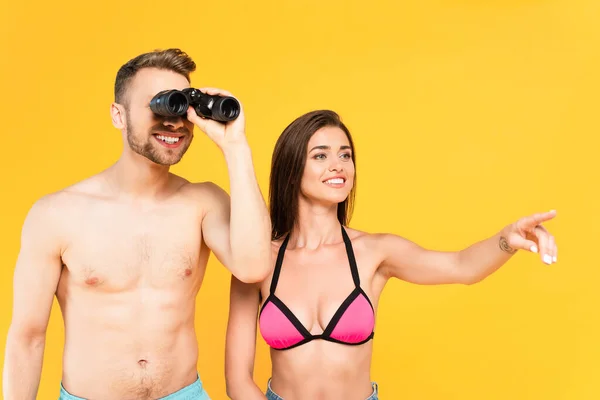 Cheerful woman pointing with finger while shirtless man looking through binoculars isolated on yellow — Stock Photo