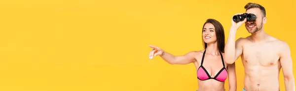 Website header of cheerful woman pointing with finger while shirtless man looking through binoculars isolated on yellow — Stock Photo