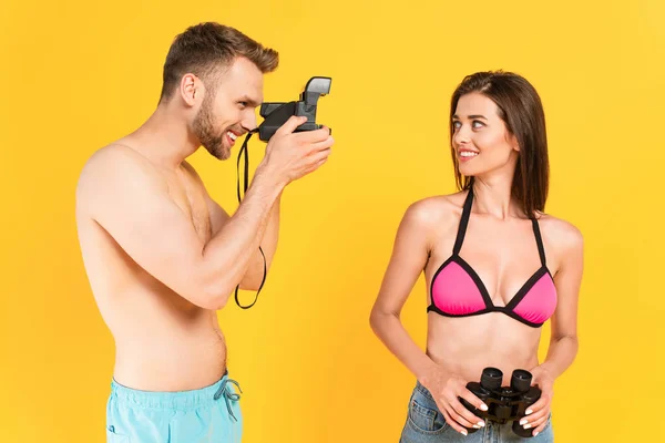 Happy man taking photo of girl in swimsuit holding binoculars isolated on yellow — Stock Photo
