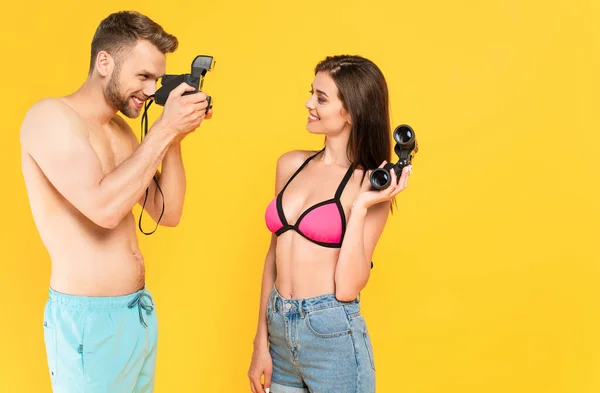 Cheerful man taking photo of girl in swimsuit holding binoculars isolated on yellow — Stock Photo