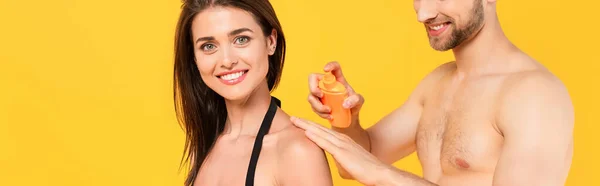 Panoramic crop of muscular man smiling while applying sunblock on cheerful woman isolated on yellow — Stock Photo