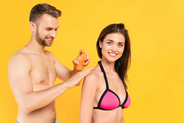 Muskulöser Mann lächelt beim Auftragen von Sonnencreme auf fröhliche Frau isoliert auf gelb — Stockfoto