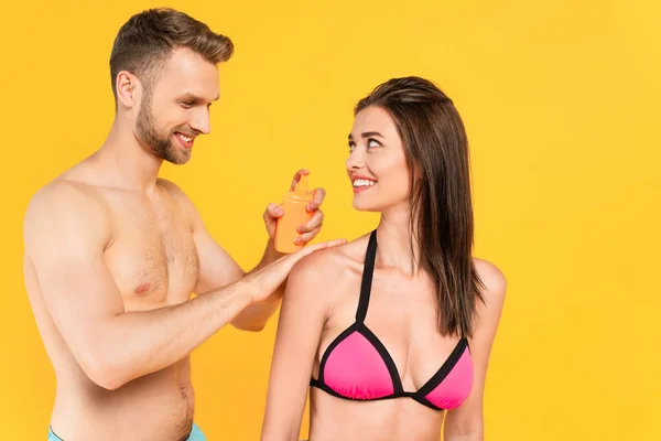 Uomo senza maglietta sorridente mentre applica la crema solare sulla donna allegra isolata sul giallo — Foto stock