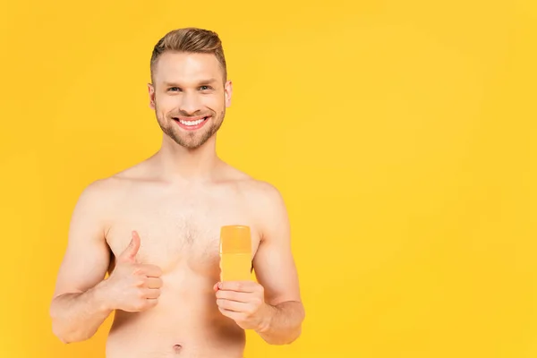 Felice uomo muscoloso mostrando pollice in su e tenendo bottiglia con crema solare isolata sul giallo — Foto stock