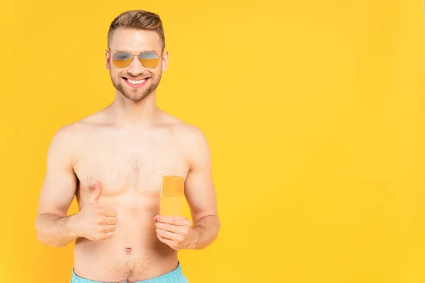 Happy muscular man in sunglasses showing thumb up and holding bottle with sunblock isolated on yellow — Stock Photo