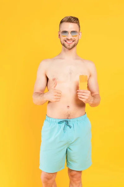 Hombre feliz sin camisa en gafas de sol mostrando el pulgar hacia arriba y sosteniendo la botella con protector solar aislado en amarillo - foto de stock
