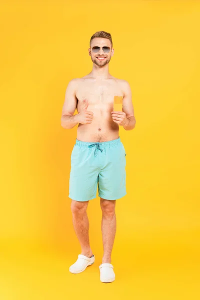 Glücklich hemdloser Mann mit Sonnenbrille zeigt Daumen hoch und hält Flasche mit Sonnencreme auf gelb — Stockfoto