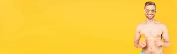 Concept panoramique d'homme heureux torse nu dans des lunettes de soleil montrant pouce vers le haut et tenant bouteille avec crème solaire isolé sur jaune — Photo de stock